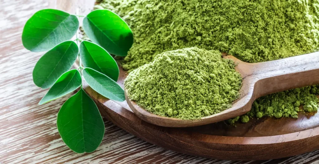 Moringa leaf powder in a spoon with fresh moringa leaves scattered on a wooden table.